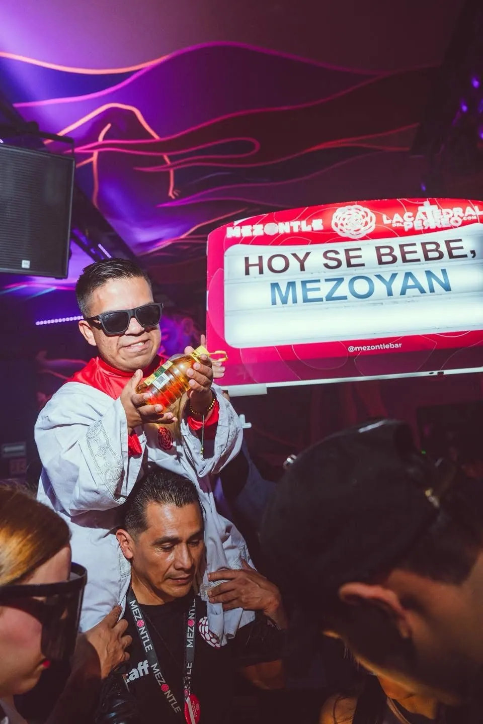 Man in sunglasses holding a bottle in front of a neon sign